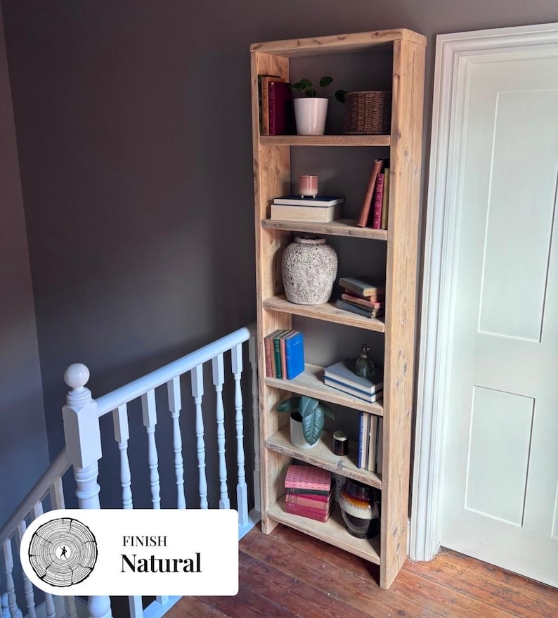 May include: A tall, narrow, light-colored wooden bookshelf with six open shelves. The shelves are filled with various items including books, a white potted plant, a woven basket, a textured ceramic vase, and a small glass bottle.