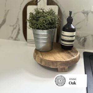May include: A small, round, wooden oak serving tray with three short legs. A small metal bucket with a green plant sits on the tray, along with a dark brown bottle with a pump. The tray has a natural wood grain finish.