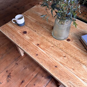 May include: A rectangular wooden coffee table with a natural light brown finish, visible wood grain, and dark knots. On the surface sits a two-toned white and dark grey ceramic mug, a clear ribbed glass vase with green foliage, and a stack of books.