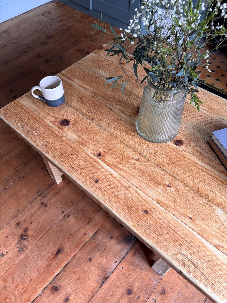 May include: A rectangular wooden coffee table with a natural light brown finish, visible wood grain, and dark knots. On the surface sits a two-toned white and dark grey ceramic mug, a clear ribbed glass vase with green foliage, and a stack of books.