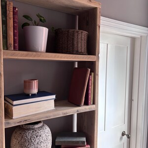 May include: A tall, light-toned wooden bookshelf with four shelves holding various items. The shelves contain stacks of hardcover books, a small potted green plant in a white ceramic pot, a woven brown basket, a pink ceramic cup, and a large textured vase.