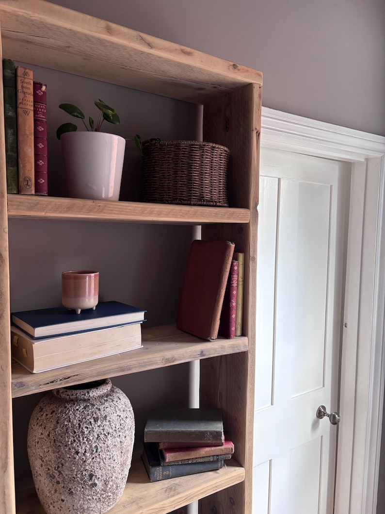 May include: A tall, light-toned wooden bookshelf with four shelves holding various items. The shelves contain stacks of hardcover books, a small potted green plant in a white ceramic pot, a woven brown basket, a pink ceramic cup, and a large textured vase.