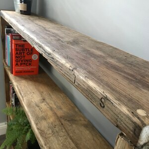May include: A tall, multi-tiered wooden bookshelf constructed from weathered, reclaimed timber with a rough, natural grain texture. The shelves hold various books, including one with an orange cover titled The Subtle Art of Not Giving a F*ck.