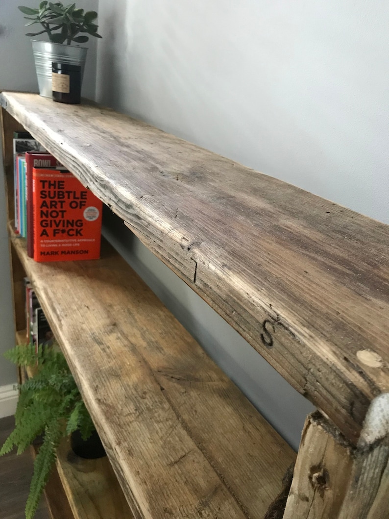 May include: A tall, multi-tiered wooden bookshelf constructed from weathered, reclaimed timber with a rough, natural grain texture. The shelves hold various books, including one with an orange cover titled The Subtle Art of Not Giving a F*ck.