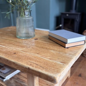 May include: A rectangular wooden coffee table with a natural light brown finish and visible wood grain. On the table sits a clear ribbed glass vase containing green foliage and a stack of two books with grey and tan covers.