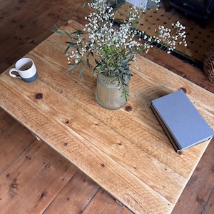 May include: A rectangular coffee table made of light brown wood with a visible grain and knots. On the table sits a clear ribbed glass vase filled with white baby's breath flowers, a two-toned white and grey ceramic mug, and a closed grey book.