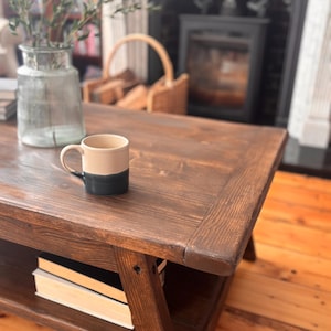 May include: A dark brown wooden coffee table with a lower shelf holding books. A two-tone beige and dark blue mug sits on the table. A glass vase with greenery and a wicker basket are in the background.