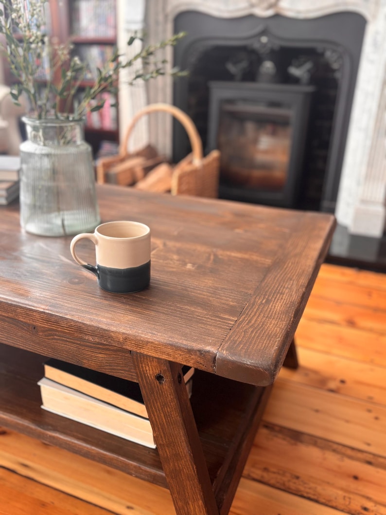 May include: A dark brown wooden coffee table with a lower shelf holding books. A two-tone beige and dark blue mug sits on the table. A glass vase with greenery and a wicker basket are in the background.