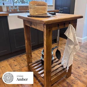 May include: A rustic wooden kitchen island with a warm amber finish, featuring a top surface holding a loaf of bread on a board, a lower slatted shelf with a small pot, and side hooks holding a black kitchen utensil and a white cloth towel.