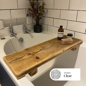 Puede incluir: Una bandeja de bañera de madera rústica con acabado transparente. La bandeja es de madera marrón claro y tiene un borde natural sin terminar. Está diseñada para descansar sobre la bañera y contiene una botella, un tarro y un cepillo. La bandeja tiene un acabado transparente.