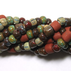 May include: Close-up of a strand of mixed beads in various colors. The beads include cylindrical rust-colored beads, and smaller round beads in shades of brown, green, and cream. The beads are strung together, creating a textured pattern.