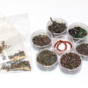 May include: An assortment of small beads in various colors and sizes. Some beads are loose in clear plastic bags, while others are contained in small, clear plastic containers. The beads appear to be metallic and iridescent, with shades of bronze, green, and red.