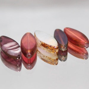 Puede incluir: Cinco cuentas de vidrio en varios tonos de rojo, rosa y blanco. Las cuentas son de forma ovalada y tienen un acabado brillante.