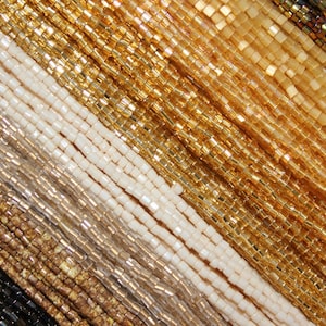 May include: A close-up of a variety of gold, beige, and white seed beads. The beads are arranged in rows and are all the same size and shape.