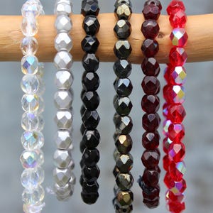 Peut inclure: Plusieurs rangées de perles à facettes de différentes couleurs sont suspendues à une cheville en bois. Les couleurs comprennent le transparent irisé, l'argent, le noir, le bronze, le rouge foncé et le rouge irisé. Les perles sont enfilées.