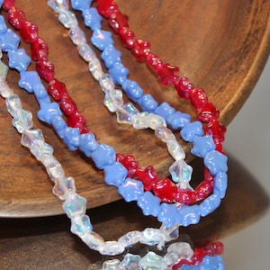 May include: Close-up of layered necklaces with star-shaped beads in red, white, and blue. The beads are strung together, creating a patriotic color scheme. The necklaces are displayed on a wooden surface.