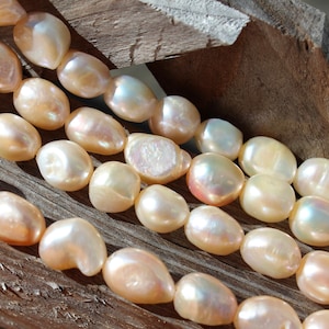 May include: A strand of off-white baroque pearls with an iridescent sheen. The pearls are oval shaped and have a slightly irregular surface. The pearls are arranged in a row on a wooden surface.