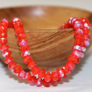 Puede incluir: Una hebra de cuentas de vidrio facetadas de color naranja brillante con brillo iridiscente. Las cuentas están enhebradas en un hilo transparente y se muestran en un cuenco de madera.