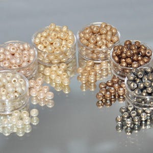 May include: An assortment of small, round beads in various colors, including cream, gold, bronze, and silver, are displayed in clear plastic containers. Some beads are scattered around the containers, reflecting on a shiny surface. These beads are ideal for jewelry making.