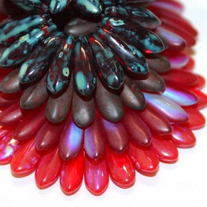 May include: Close-up of a decorative floral-shaped object. The object features layers of teardrop-shaped beads in shades of red, brown, and teal. The beads have a glossy finish and are arranged in a radial pattern, creating a flower-like appearance.