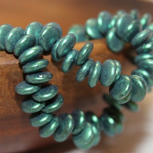 May include: A teal-colored beaded bracelet. The beads are flat, oval-shaped, and have a metallic sheen. The bracelet is resting on a wooden surface, showcasing its color and texture. This piece of jewelry is suitable for everyday wear.