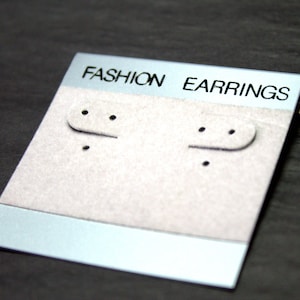 May include: A white card with a blue border and the text "FASHION EARRINGS" printed on it. The card has two holes for displaying earrings.