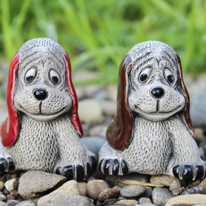 May include: Two ceramic dog figurines with big eyes and floppy ears. One dog is wearing a red hat and the other is wearing a brown hat. The dogs are sitting on a bed of small gray rocks.