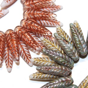 May include: Close-up of various glass beads in a leaf shape. The beads are in shades of white, copper, and gold. The leaf-shaped beads are arranged in a circular pattern, with some beads overlapping.