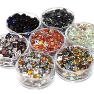 May include: An overhead shot of seven small, clear plastic containers filled with various colorful beads. The beads are in shades of black, silver, orange, green, and iridescent colors. The containers are arranged in a circular pattern.