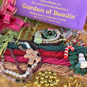 May include: A collection of Christmas-themed beads, charms, and findings in various colours, including red, green, gold, and white. The beads are arranged on a woven surface and include a gingerbread man charm, a candy cane charm, and a snowman charm. The image also includes a purple pouch with the text "Garden of Beadin'" and contact information.