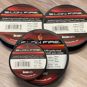May include: Three spools of S-LON FIRE braided and fused bead thread. The spools are black with red and white labels. The labels list the thread's color, size, and weight. The thread is available in Crystal, Black, and  Noir.