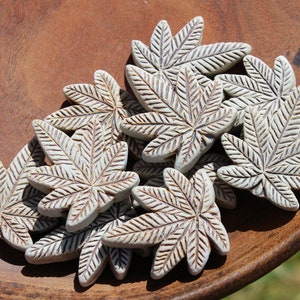 May include: A collection of ceramic leaf-shaped tiles in a light grey colour with brown veining. The tiles are arranged in a pile on a wooden surface.