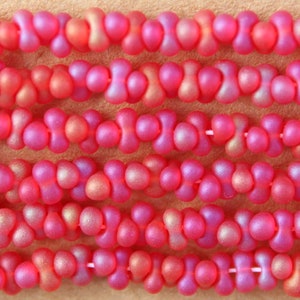 May include: Close-up of pink glass beads with a textured, iridescent finish. The beads are shaped like a double-loop or figure eight.