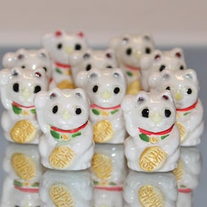 May include: A group of ten white ceramic figurines of lucky cats, also known as Maneki-Neko, with raised paws. Each cat has a red and green collar and a gold coin in its paw.