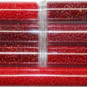 May include: A collection of red beads in various shades, stored in clear plastic tubes. The beads range from bright cherry red to deeper burgundy tones, offering a variety of textures and finishes for crafting projects. The tubes are arranged vertically.