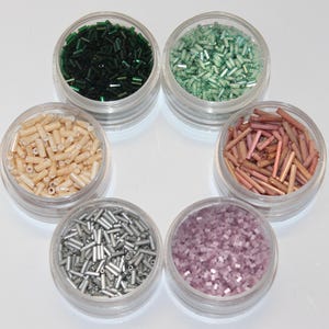May include: Six small containers filled with various colors of glass beads. The beads are in the shapes of small squares and long cylinders. The colors of the beads are green, light green, cream, silver, pink, and brown.