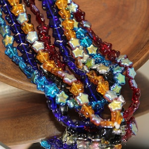 May include: Assortment of iridescent star-shaped glass beads in blue, red, orange, and gold colors. The beads are strung on a wire and are displayed in a wooden bowl.