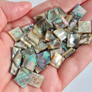 May include: A pile of iridescent abalone shell tiles in various shades of green, brown, and gold. The tiles are square shaped and have a natural, organic look.