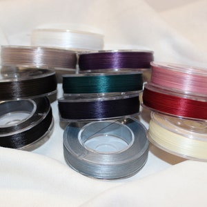 May include: Spools of thread in various colors, including black, white, gray, red, pink, purple, and green. The thread is wound around clear plastic spools.