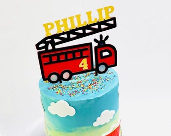May include: A blue and green ombre cake decorated with a red fire truck and the name "Phillip" on top. The cake is decorated with colorful sprinkles and clouds. There are also two toy trucks on the side of the cake.