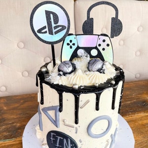 Puede incluir: Un pastel de goteo blanco y negro decorado con un mando de PlayStation gris y negro, un logotipo de PlayStation blanco y negro y unos auriculares negros. El pastel está decorado con formas geométricas grises y espolvoreado con sprinkles blanco y negro. El pastel está sobre una base de pastel blanca. El pastel dice "Fin 13" en la parte delantera.