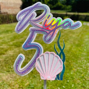 Op de afbeelding: Een paarse en iriserende taarttopper met de naam "Riya" en het nummer "5" erop. De topper heeft ook een roze schelp en groen zeewier.