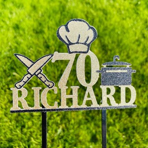 May include: A gold glitter cake topper with the name "RICHARD" and the number "70". The topper includes a chef's hat, crossed knives, and a pot. The background is green grass.
