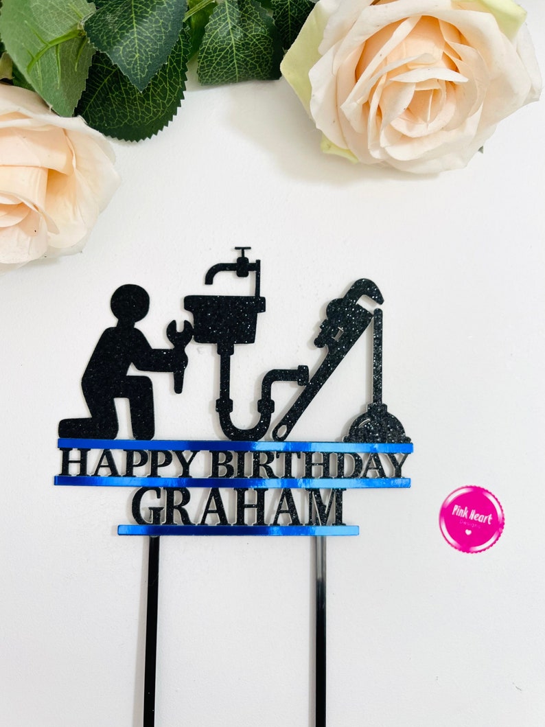 May include: Black cake topper with a silhouette of a plumber holding a wrench and a sink. The topper has a blue ribbon with the text "Happy Birthday Graham".