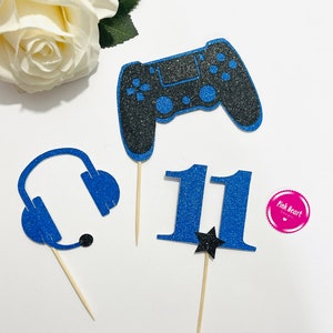 Peut inclure: Trois décorations de gâteau en paillettes bleues et noires. Une décoration est une manette de jeu vidéo, une autre est un casque audio et la dernière est le chiffre 11 avec une étoile noire.