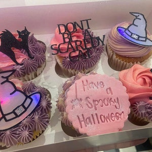 Può includere: Sei cupcake decorati con temi di Halloween. I cupcake sono glassati in rosa e viola con accenti di glitter nero e bianco. I cupcake sono sormontati da varie decorazioni a tema Halloween, tra cui un cappello da strega, un gatto nero e un cartello che dice "Non essere un fifone" e "Buon Halloween spaventoso".