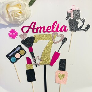Puede incluir: Conjunto de adornos para tartas con un nombre "Amelia" de purpurina rosa, un número 7 de purpurina dorada, un corazón de purpurina rosa y dorada, un pincel de maquillaje de purpurina negra y rosa, una máscara de pestañas de purpurina rosa y dorada, un pintalabios de purpurina negra y rosa, un frasco de esmalte de uñas de purpurina rosa y dorada y una silueta negra de purpurina de una niña con un lazo rosa.