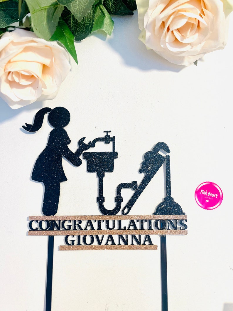 May include: Black glitter cake topper with a silhouette of a woman holding a wrench and a sink. The topper also includes a plunger and a pipe. The text "CONGRATULATIONS GIOVANNA" is on a gold glitter banner.