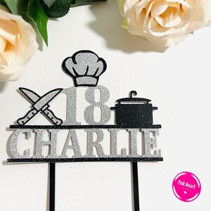 May include: A black and silver glitter cake topper with the name "Charlie" and the number "18". The topper features a chef's hat, crossed knives, and a cooking pot. The background includes roses.
