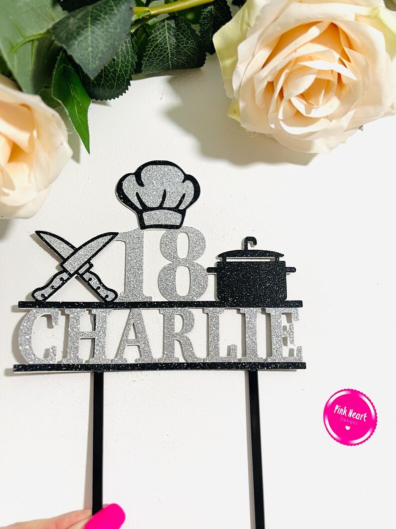 May include: A black and silver glitter cake topper with the name "Charlie" and the number "18". The topper features a chef's hat, crossed knives, and a cooking pot. The background includes roses.
